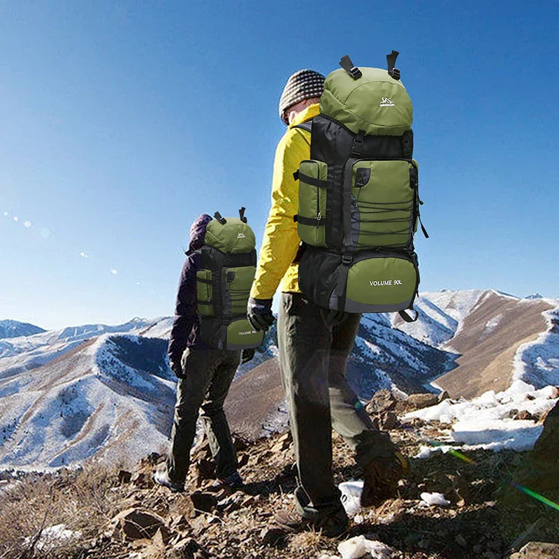Two hikers with green backpacks standing on a mountain with snow-capped peaks.